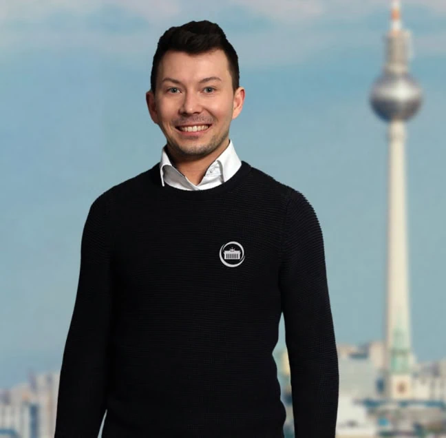 Mann mit dunklem Pullover und weißem Hemd, lächelnd vor einem verschwommenen Hintergrund mit dem Fernsehturm in Berlin.