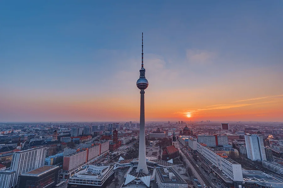 Fernsehturm in Berlin während eines Sonnenuntergangs mit orangefarbenem Himmel und Stadtansicht im Hintergrund.
